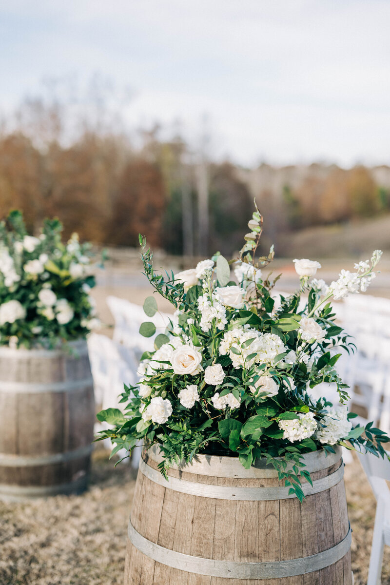 Wooden barrels with white floral arrangements sitting on top outside with aisle of white chairs