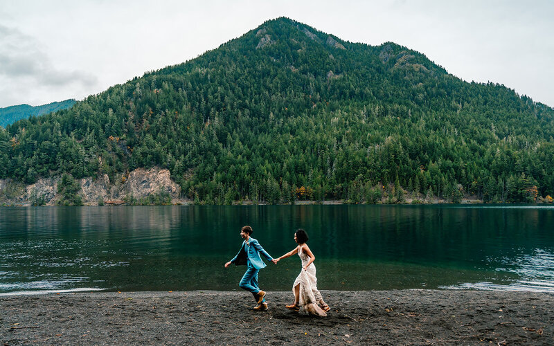 during their sunset elopement package, we see two brides in their wedding attire running a long the shore of a lake crescent. An emerald green mountain can be seen perfectly centered behind them. 