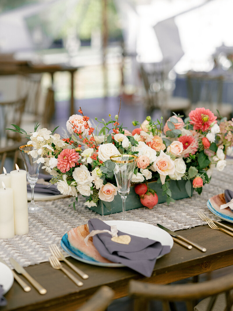 Close of up wedding reception table with white and coral red flower centrepiece, gold cutlery, and painted coral and teal plates, with a grey serviette.