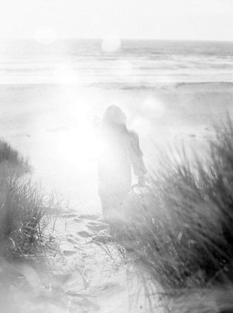 Black and white shot of a woman walking past sand dunes to the ocean, meant to represent a past life trauma.