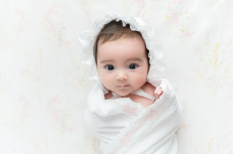 Newborn baby with brown hair and eyes wide open in a muslin swaddle and heirloom bonnet