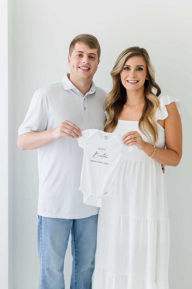 Woman and man holding baby onesie to announce pregnancy during a photoshoot in Dothan Alabama Photography Studio