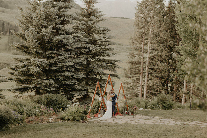 There's a wedding couple in front of a tringle style altar cuddling for a portrait. With a Mountain scene behind them.