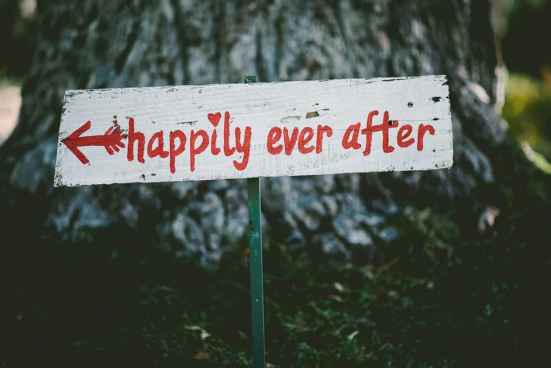 Rustic wooden sign with red lettering that reads 'happily ever after' and an arrow pointing left, placed outdoors in front of a large tree trunk