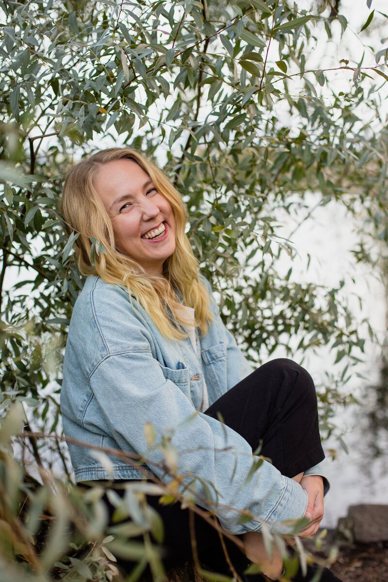 mielen paremminvoinnin ja kehomieliyhteyden valmentaja  hymyilee  ja istuu puunrungon päällä puunoksien keskellä