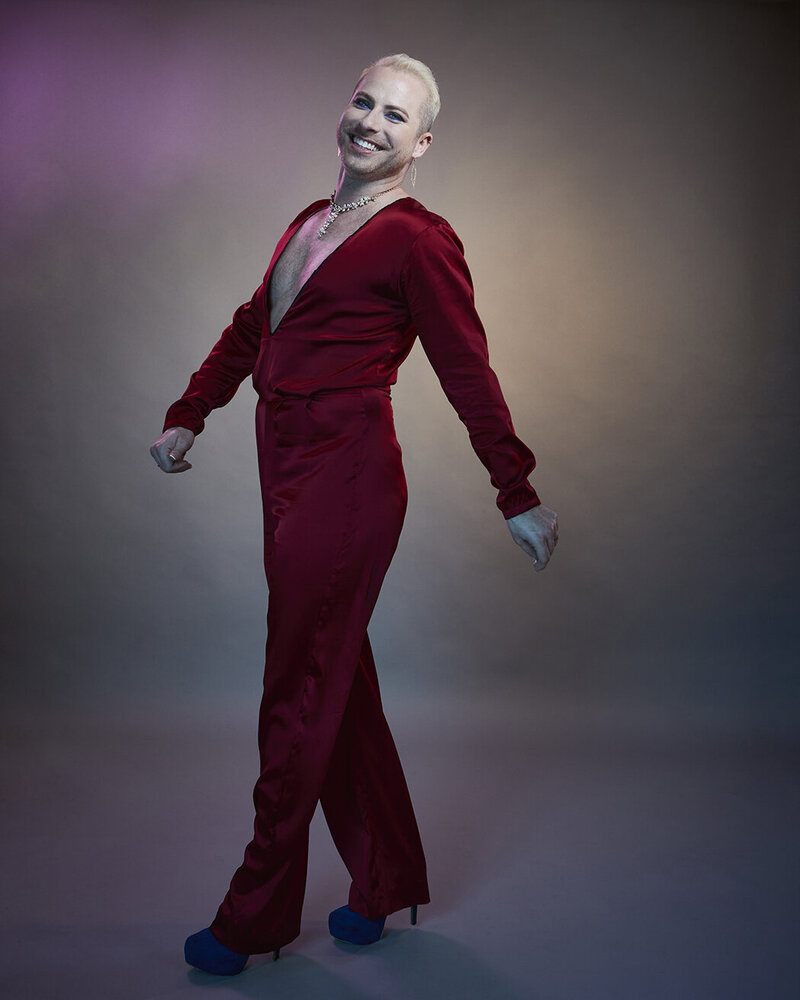 Stylized full-body portrait of Dain August in a red ensemble, striking a dynamic pose against a dramatic background, captured by Ylva Erevall.
