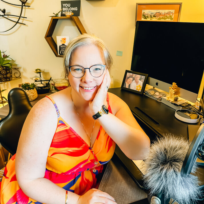 This is a photo of a smiling Sandra Booker, with glasses, seated at a desk with a computer and various desk items. She has blonde hair pulled back and is wearing a colorful dress with a v-neck. There are framed pictures, a plant, and decorative items in the background, as well as a sign that says "YOU BELONG HERE." The environment suggests a home office setting. The image is well-lit and conveys a friendly, welcoming atmosphere.