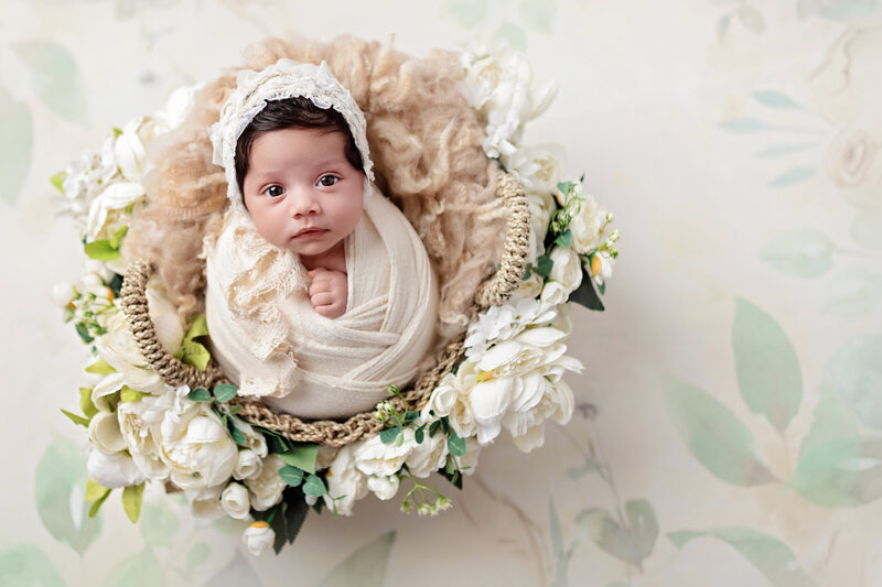 Newborn baby girl wrapped in cream, sitting in a floral basket with white flowers and soft textured wool, looking up with bright eyes.