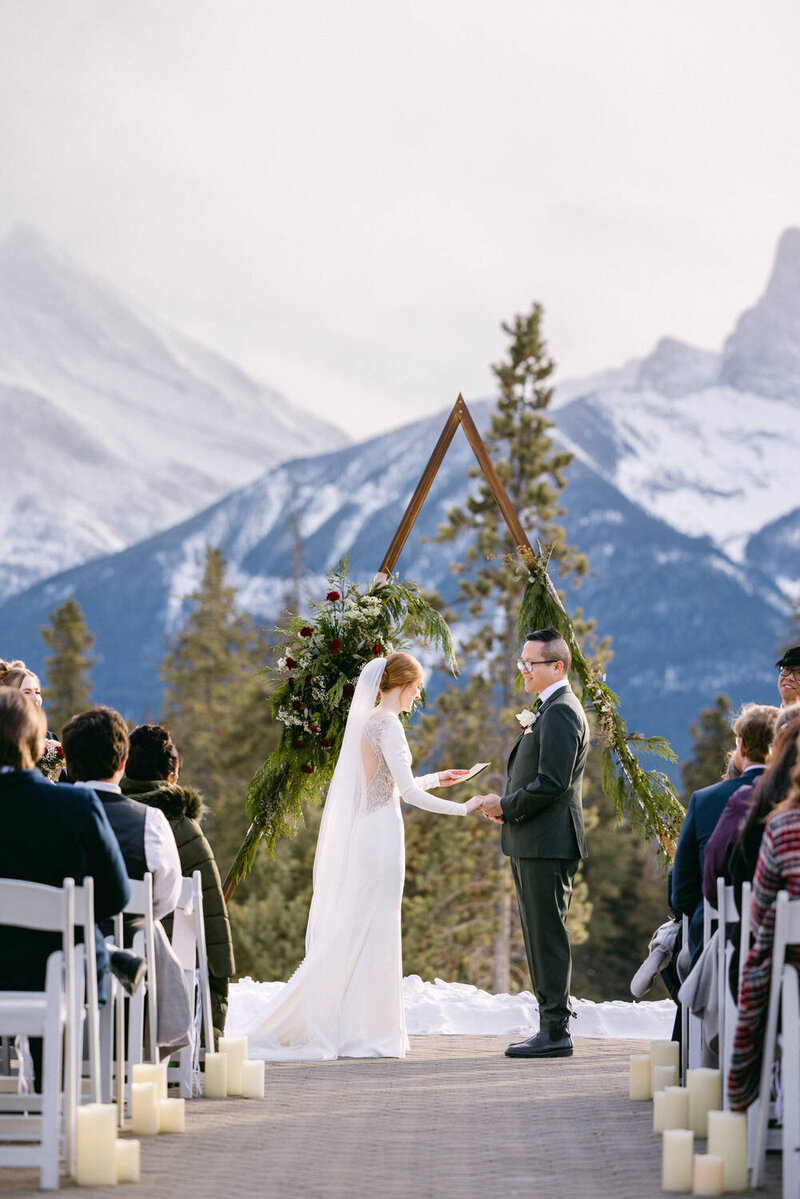 The bride and groom exchange vows in a beautiful winter ceremony at Silvertip Golf Course in Canmore, with the iconic Three Sisters mountains creating a breathtaking snowy backdrop.