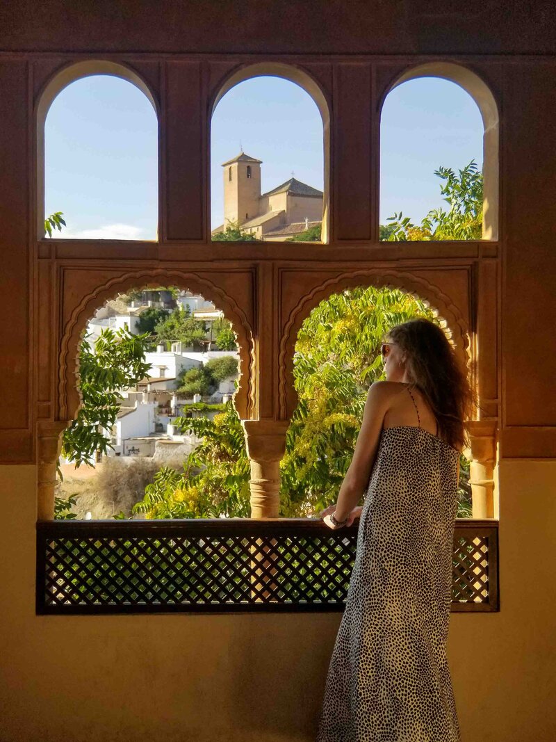 Award-winning luxury travel expert Stephanie Dosch admiring the view of whitewashed buildings and a historic church in Granada, Spain, through a Moorish-style window ©Stephanie Dosch | theViatrix Luxury Travel