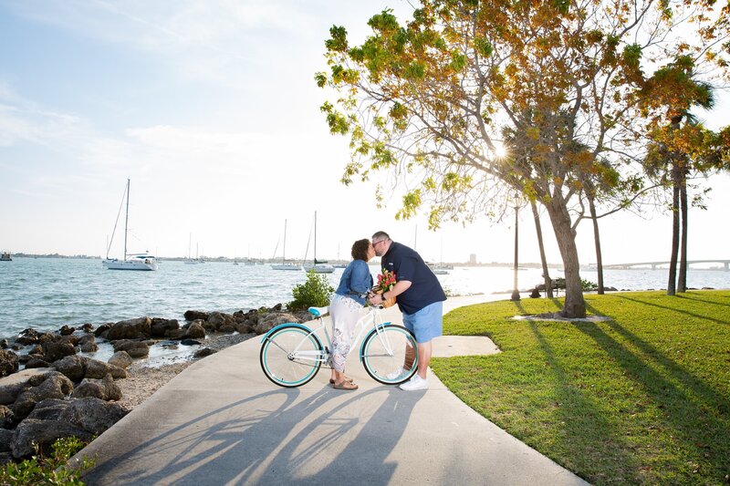 Engaged couple kissing on the sidewalk path at Bayfront Park in Sarasota with sailboats in the background