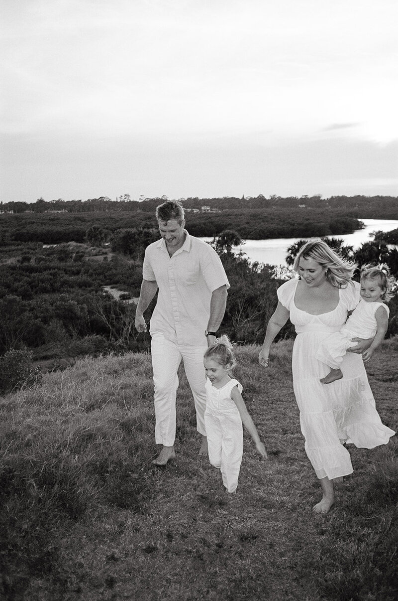 Black and white photo of a barefoot family walking up a grassy hill during a maternity shoot in New Smyrna Beach.