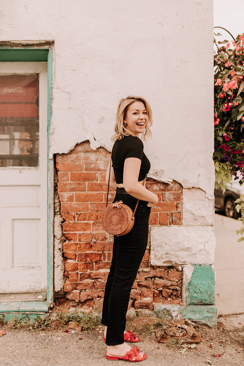 Lauren Hughes is standing next to a brick wall and floral hanging basket smiling. She's wearing all black, red shoes and a circular basket bag.