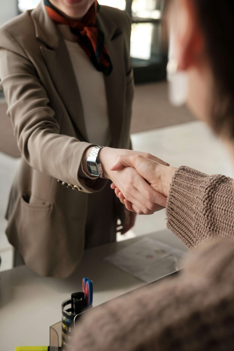 Two people shaking hands in a professional setting, one wearing a tan blazer and scarf, and the other in a knitted sweater, with office supplies visible in the background.