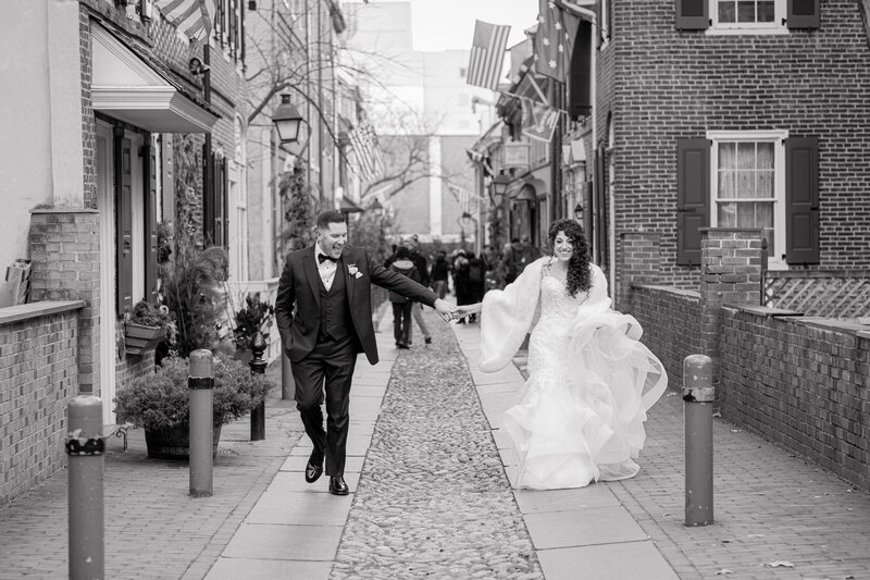 Bride and groom walking hand in hand down the historic Elfreth's Alley in Philadelphia, captured in black and white.