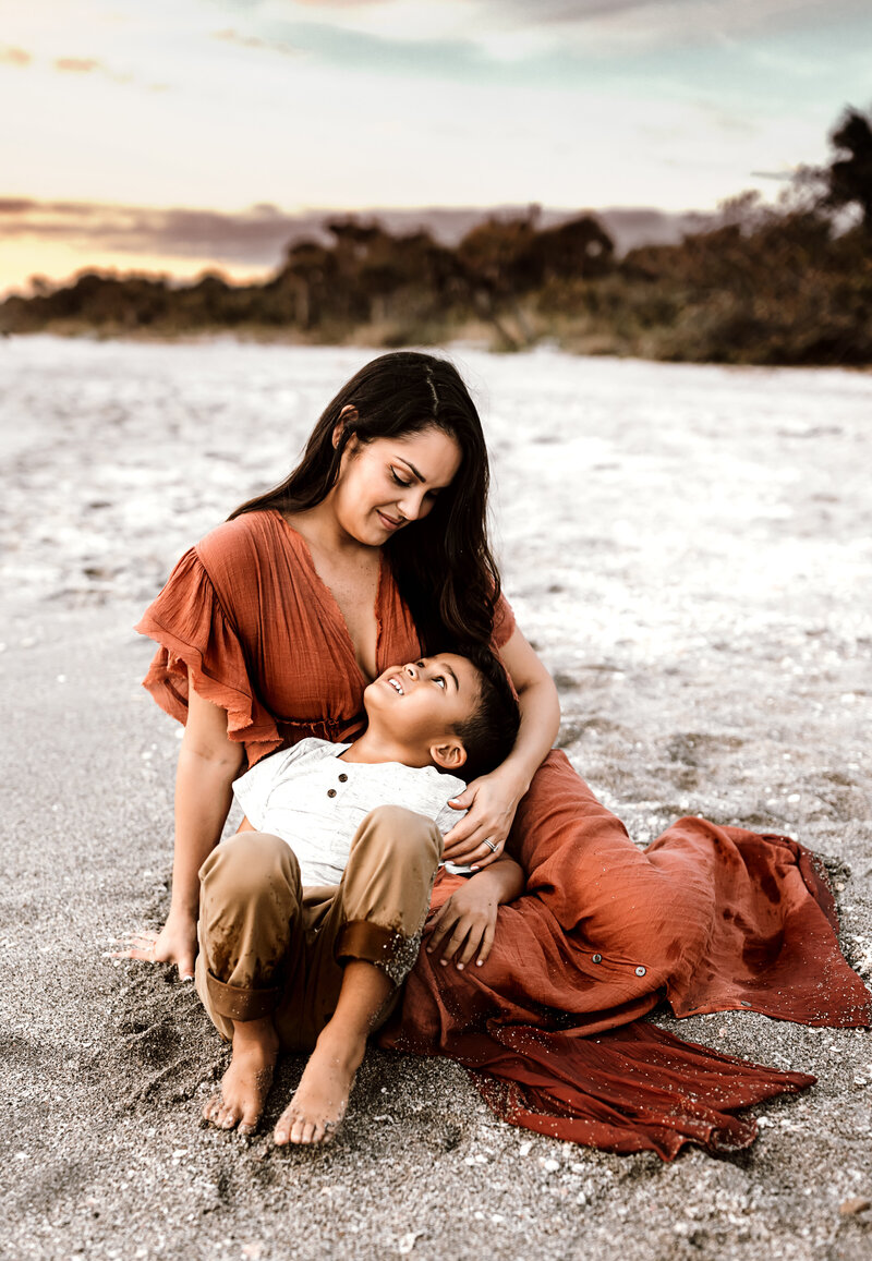 Family Photographer, a young mother looks gazes back at her young son on her lap at the beach