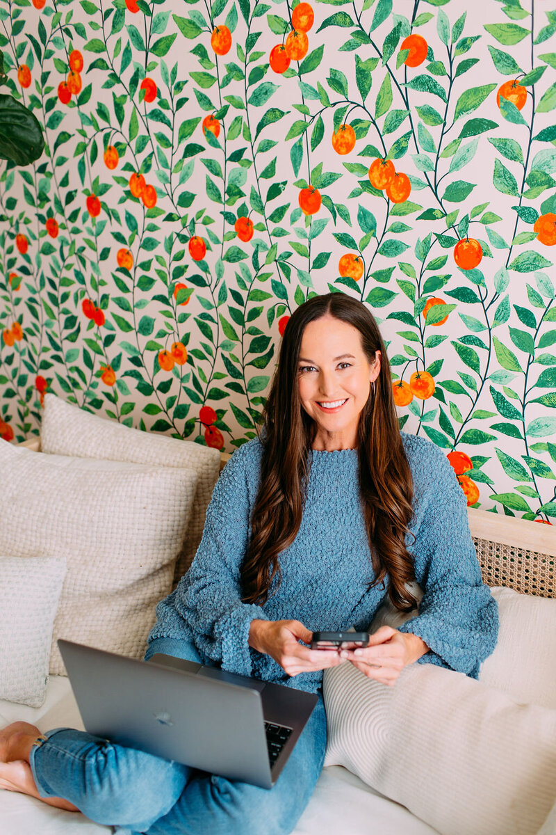 Kristie wearing a blue sweater, sitting on a couch in front of a colorful wall, smiling for the camera.