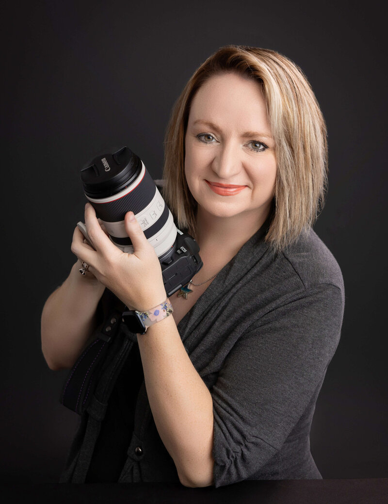 Christy Whitehead, photographer, in dark grey on a black background holding a Canon mirrorless camera.