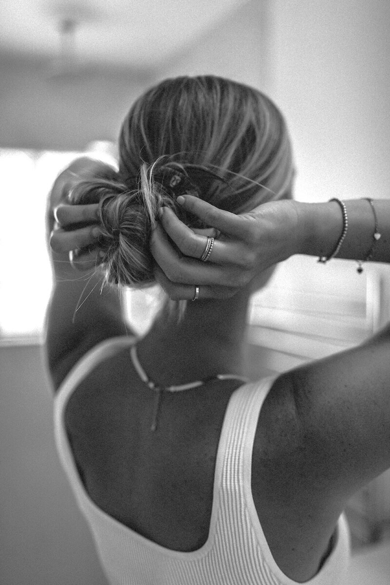 A black and white photograph captures a woman from behind, dressed in a white top, as she gathers her blonde hair into a casual bun with an air of quiet elegance.