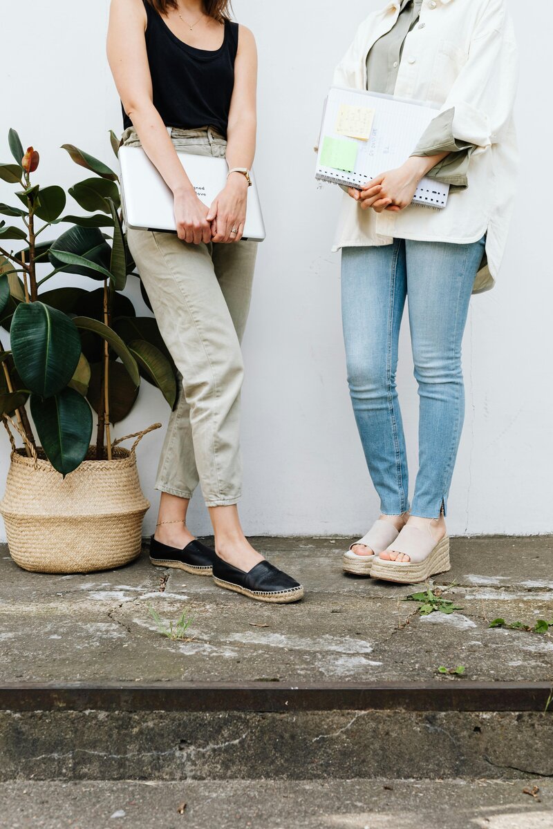 two woman in casual clothing standing with a laptop and notebook
