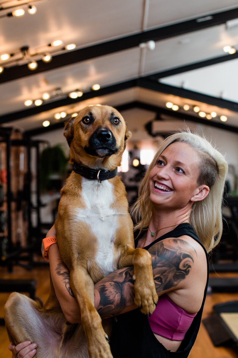 A woman with tattoos and a bright smile holding a large brown dog in a dog-friendly Melbourne gym, illustrating the gym's inclusive and friendly atmosphere.