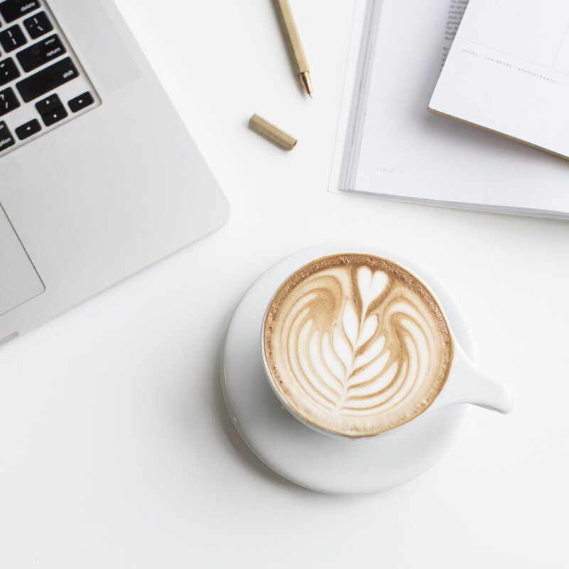 A mug with latte art next to notebooks and a laptop