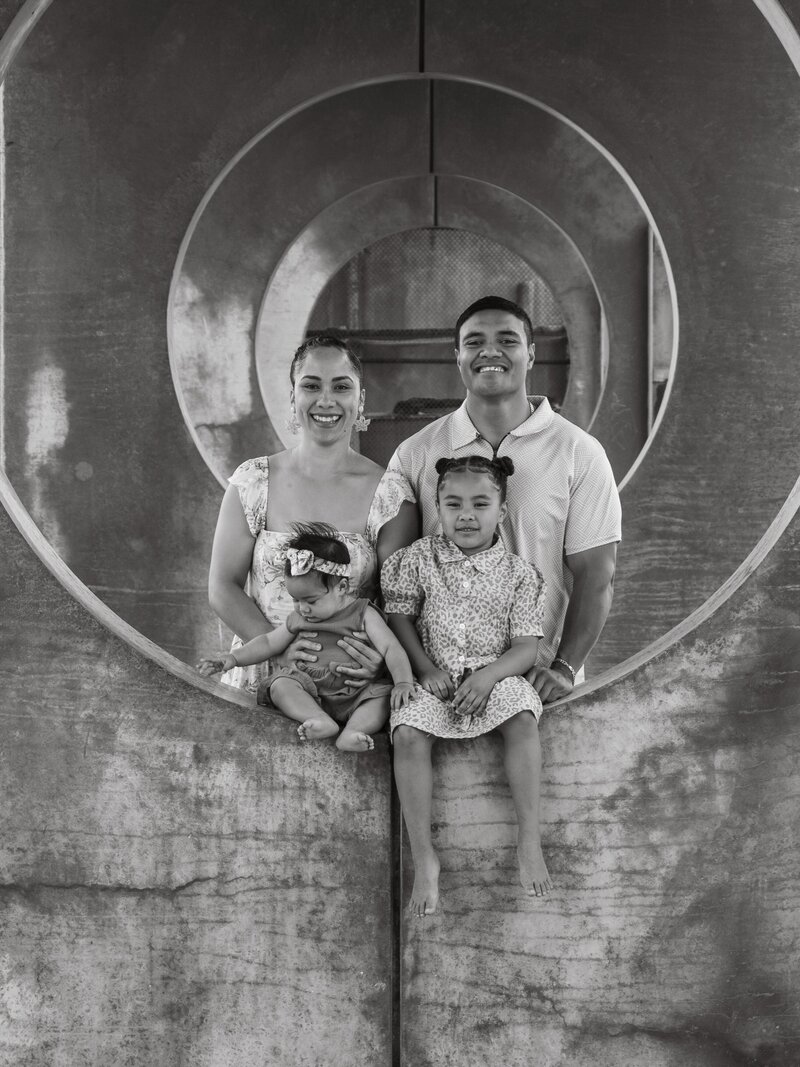 black and white family portrait with mom, dad, and young daughter smiling at the camera, plus baby girl playing and being held by her mother. they are at New Brighton Pier in Christchurch, New Zealand in summer.