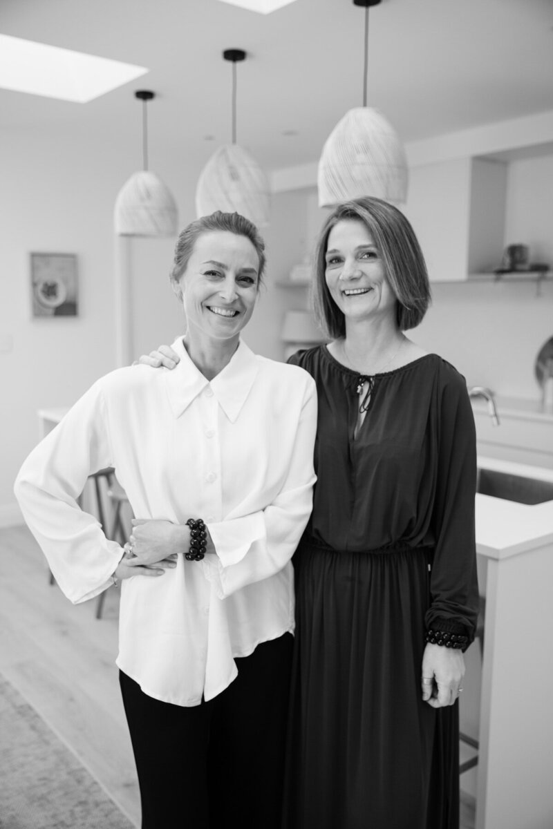 Naturopath Lauren stands on the left, with spiritual healer Sharon on the right. Both are in a modern kitchen setting, black and white photo.