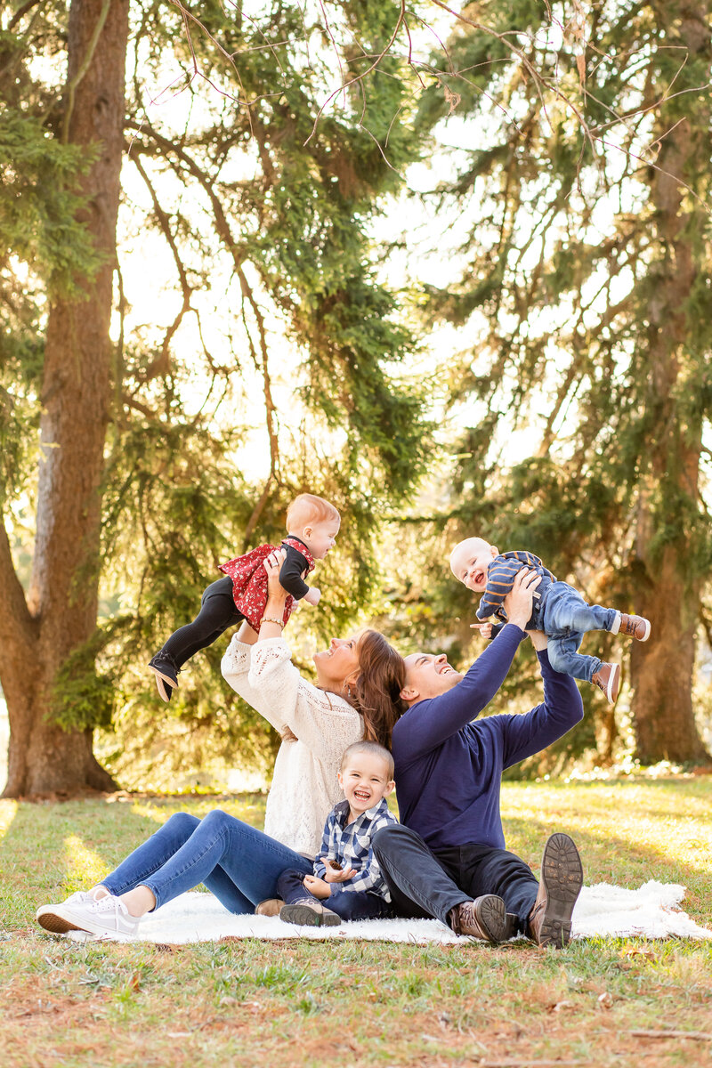 MOm and dad throwing twin babies up sitting on blanket with toddler laughing with a pine tree backdrop