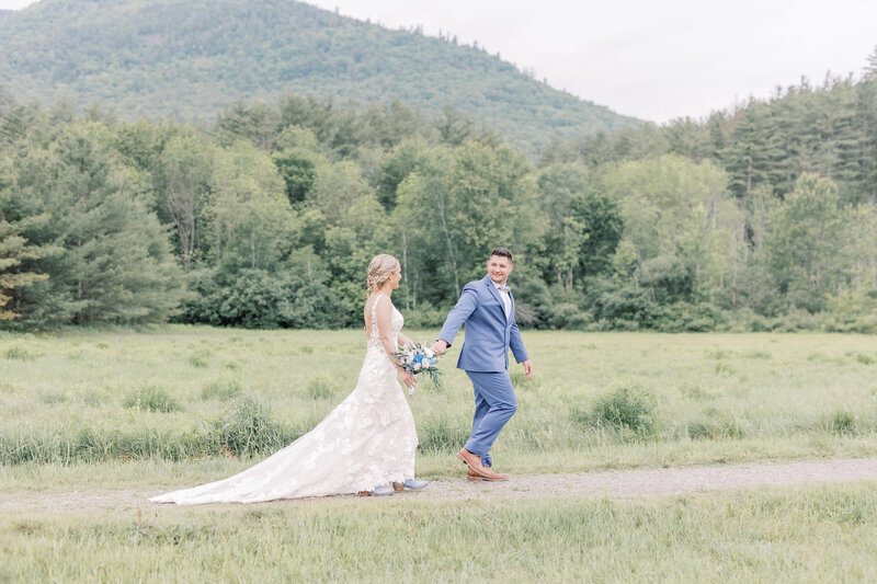 On a dirt path in Keene Valley with the Adirondack mountains behind them, a bride and groom are holding hands and walking together after their ceremony during their elopement.