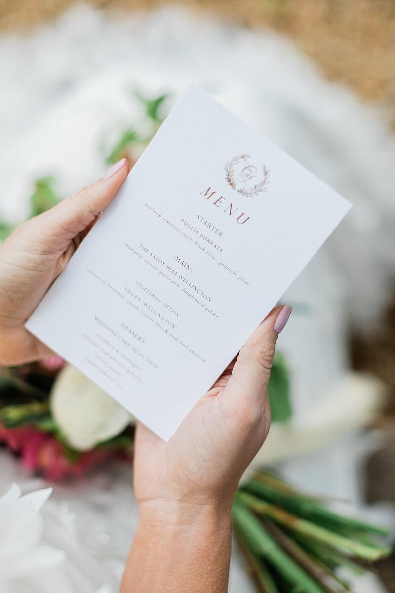 Elegant printed wedding menu in blush and burgundy, held by the bride with a bouquet in the background