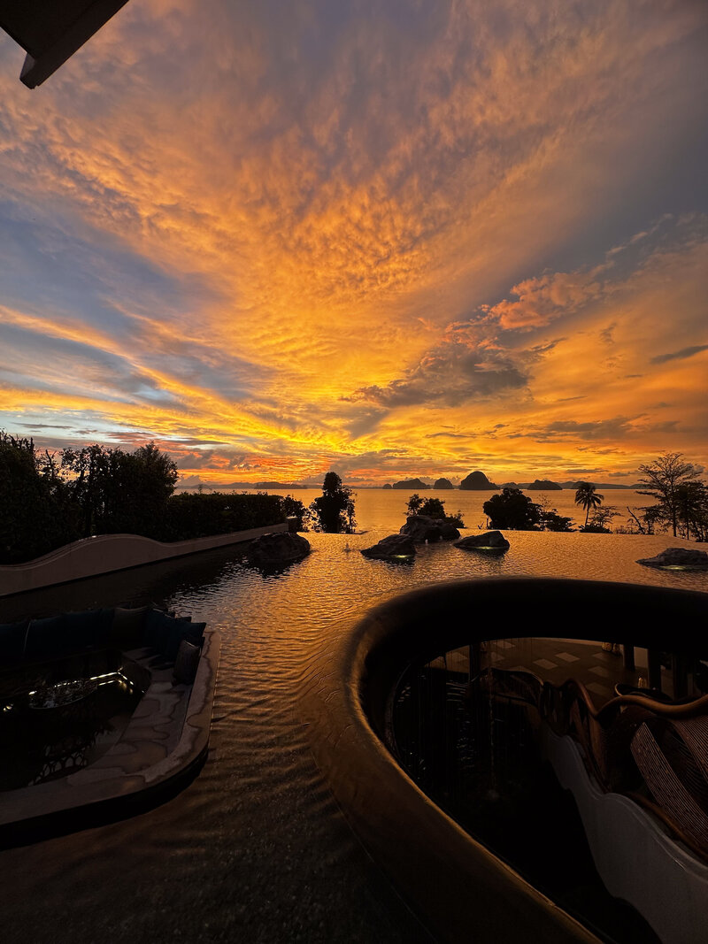 A breathtaking sunset view with the sky painted in orange, pink, and purple hues over a body of water. The silhouettes of trees and small islands are visible in the distance, creating a serene and dramatic scene.