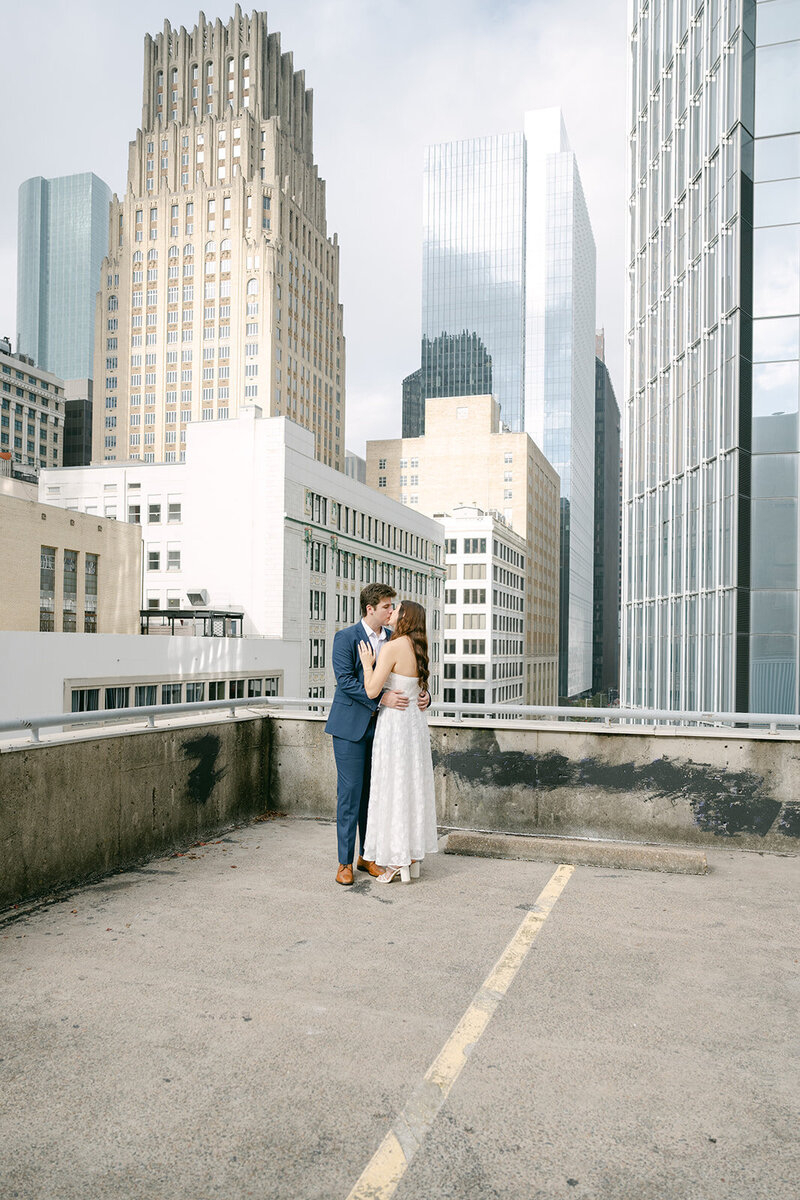 Bride and groom share a kiss on a rooftop in downtown Houston with the skyline behind them, captured in an editorial fine art style by a Texas destination wedding photographer.