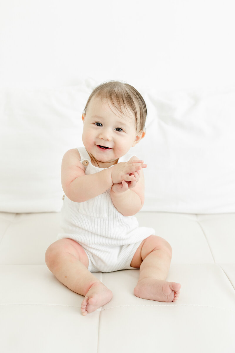 Baby boy sits in white overalls and smiles during portrait session