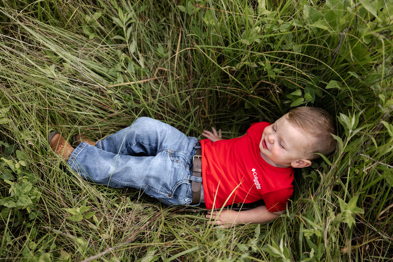 Toddler boy wearing blue jeans and red shirt laying in tall grass smiling. 
