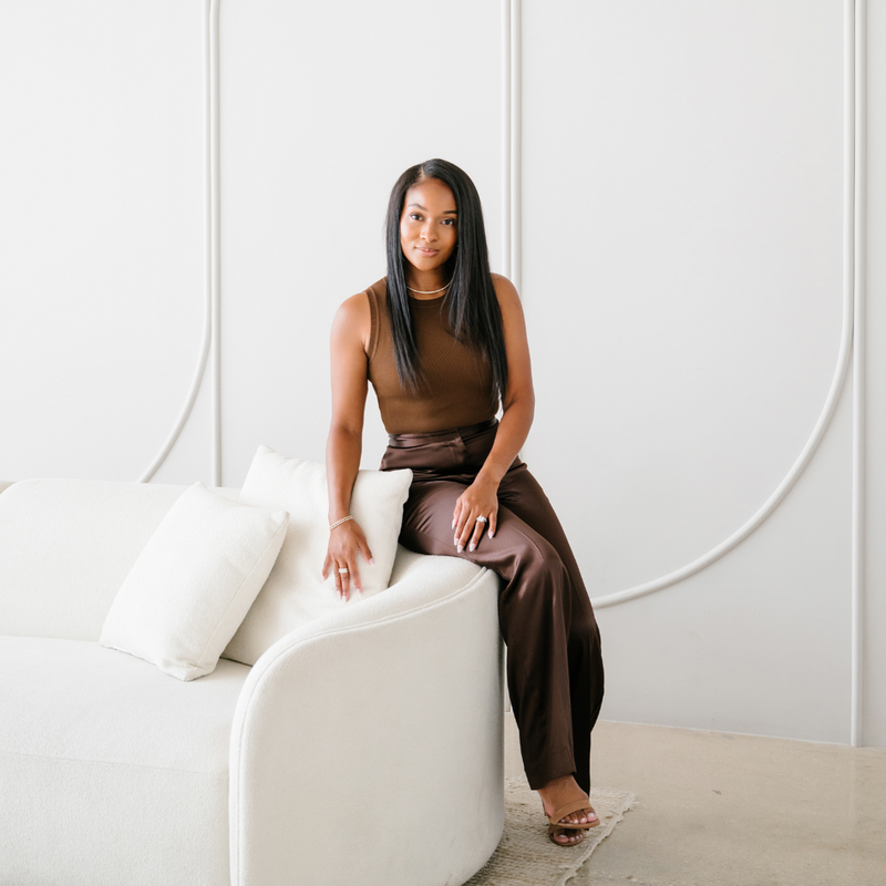 Courtney Alberino, co-founder of Interiors by CV, sitting on a light sofa in a modern living room, dressed in a sleeveless top and dark pants.