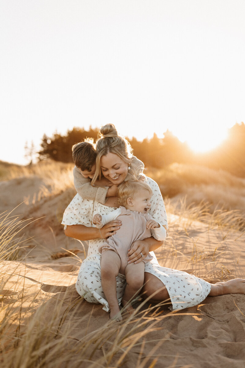 A mother being hugged by her baby and son while the sunsets in the treeline behind them and posing for the camera during a family photoshoot in christchurch with lucy jane photography