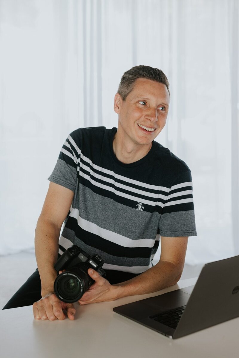 Photo of Chris Perkins, Photographer at Underatreehouse Photography, a commercial event photographer on the Central Coast. The image shows him sitting behind a laptop at a desk with his camera in his hand. He is smiling and looks friendly.