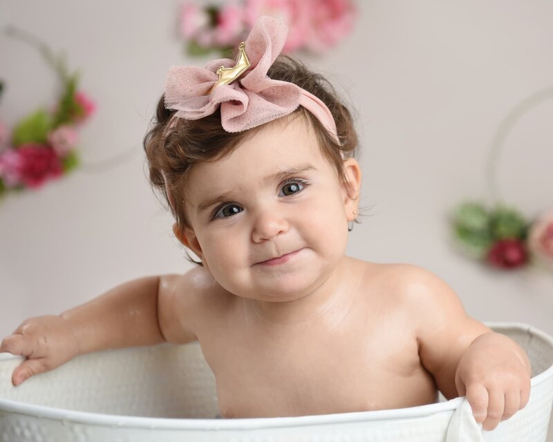 A baby girl with a pink bow headband sitting in a white tub, surrounded by pink floral decorations, smiling at the camera.