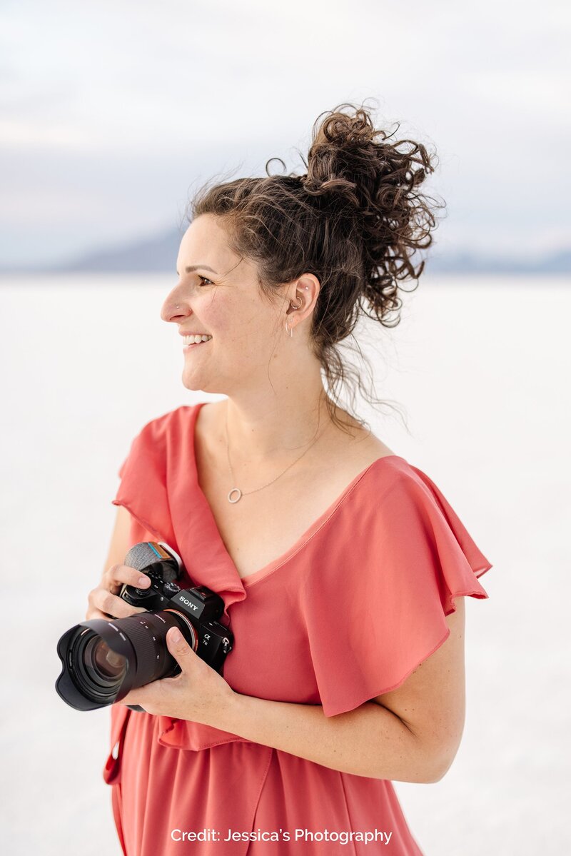 Kate Simpson stands outdoors holding a camera, smiling as she looks off into the distance. She wears a coral-colored dress and her curly hair is styled in a loose bun.