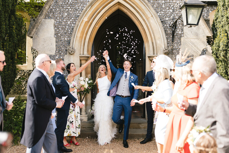 A joyful bride and groom exit a stone church, holding hands and celebrating. Friends and family surround them, tossing flower petals. The scene is filled with smiles and laughter, capturing a happy wedding moment on a sunny day.