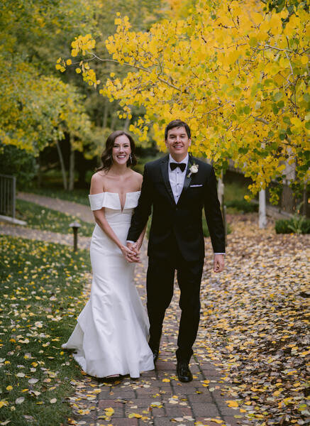 Bride and groom hold hands outdoors in the fall with yellow leaves.