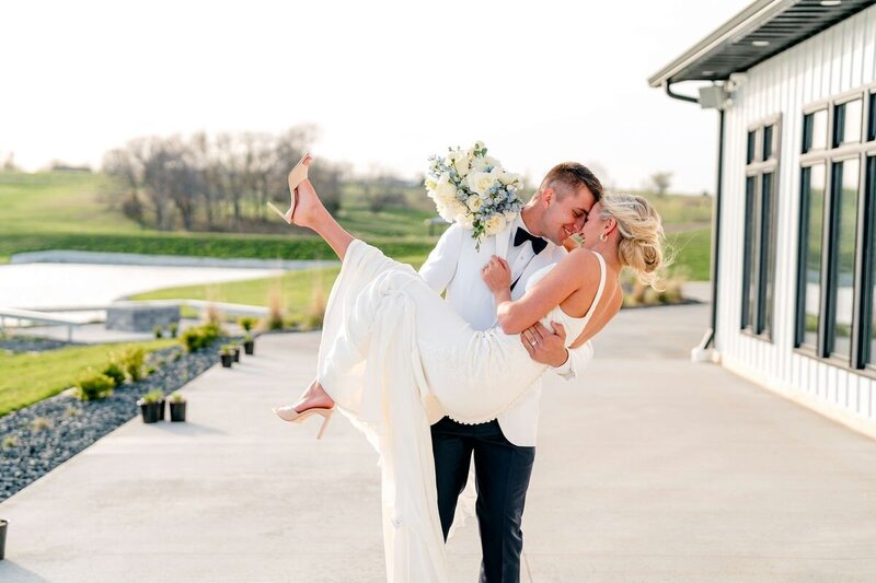 A groom holding his new bride in the air sharing an intimate moment smiling at each other outside in front of a lake.