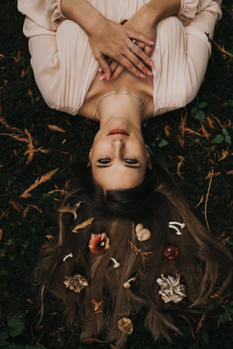 Woman with long, brown hair lays on forest floor with her hands on her chest as she looks at the camera. Small mushrooms and foliage are laid flat across her outspread hair.