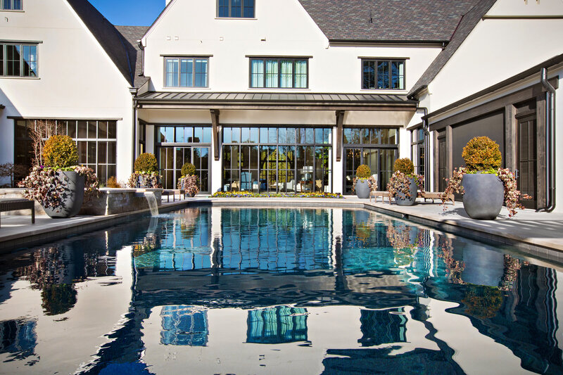 Modern home with large windows and a reflective pool, surrounded by potted plants and minimalist outdoor landscaping.