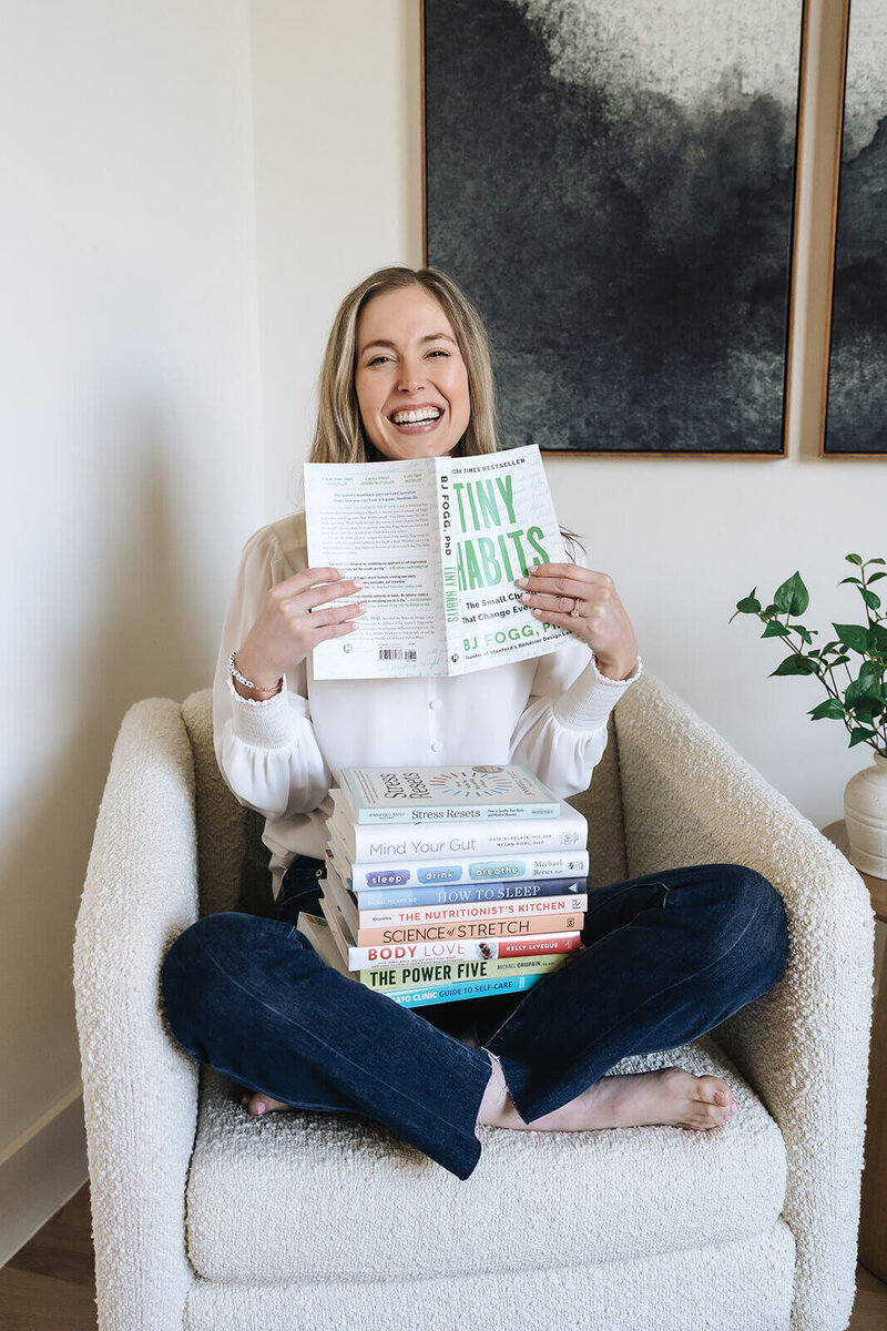 Erica Taylor, certified health and wellness coach for moms, smiling with a stack of wellness books that inspire her real-life approach
