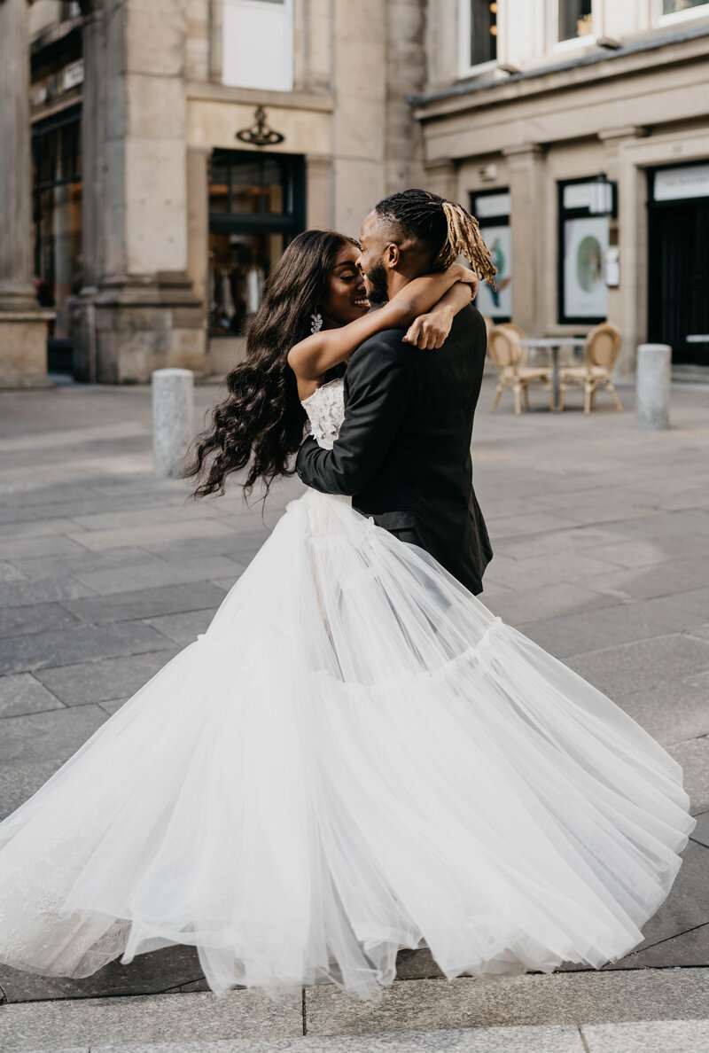 A close photography of a groom as he spins his bride around in the air in a Glasgow city square after their wedding. Her wedding dress floats in the air.