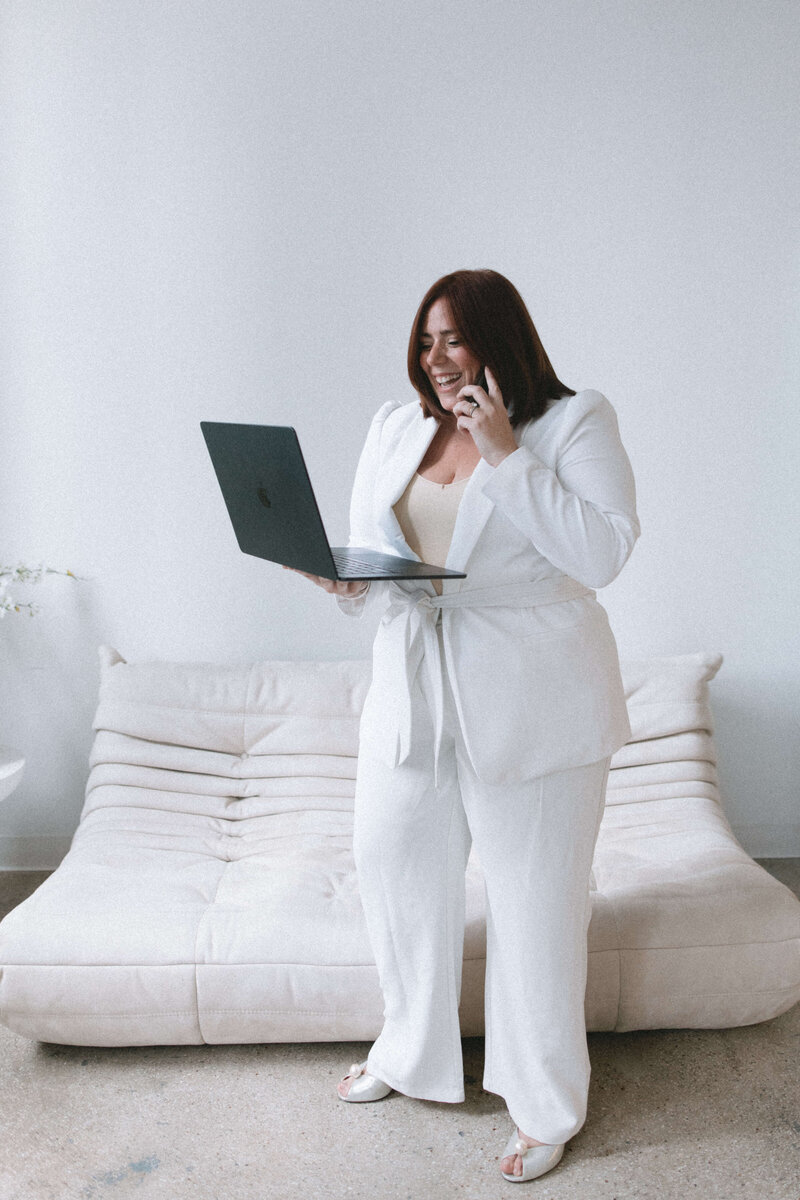 Casey, Founder of Most Valuable Bridesmaid laughing on the phone and holding laptop wearing a white suit.