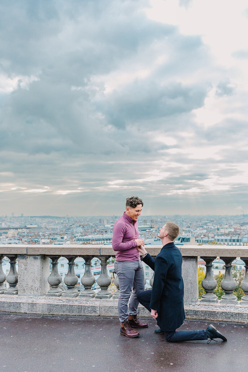 Gay couple during a Paris proposal at Sacré-Cœur in Montmartre — one man kneeling as the other joyfully accepts.