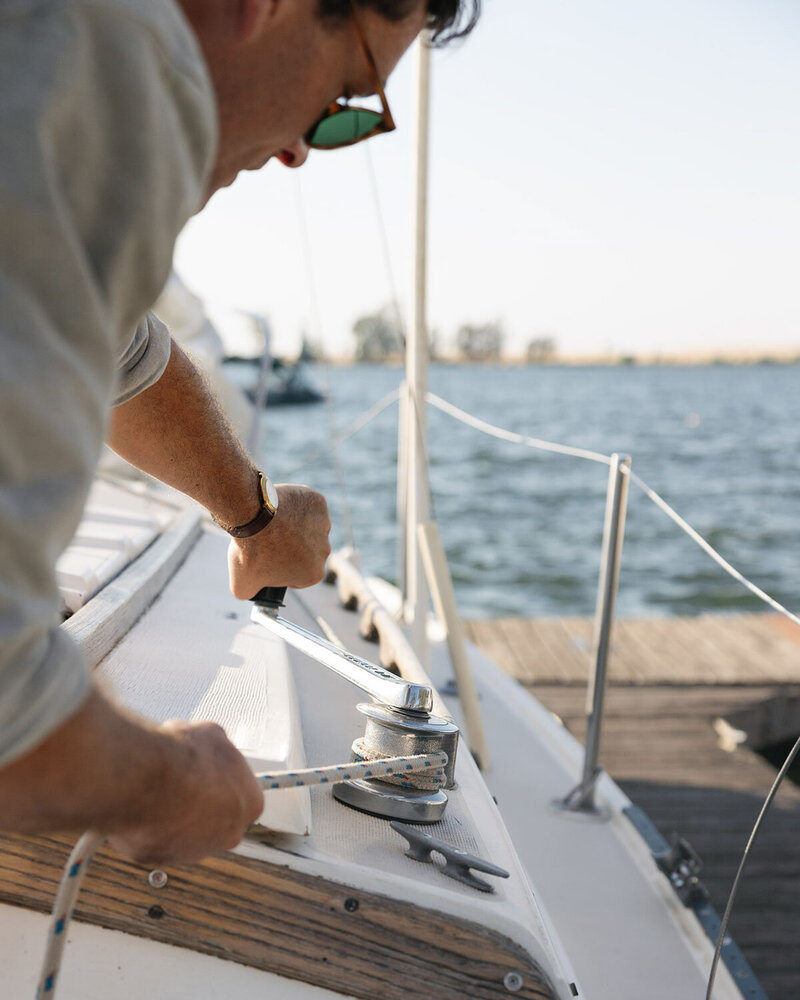 A person wearing sunglasses and a watch is adjusting a rope on the deck of a sailboat.